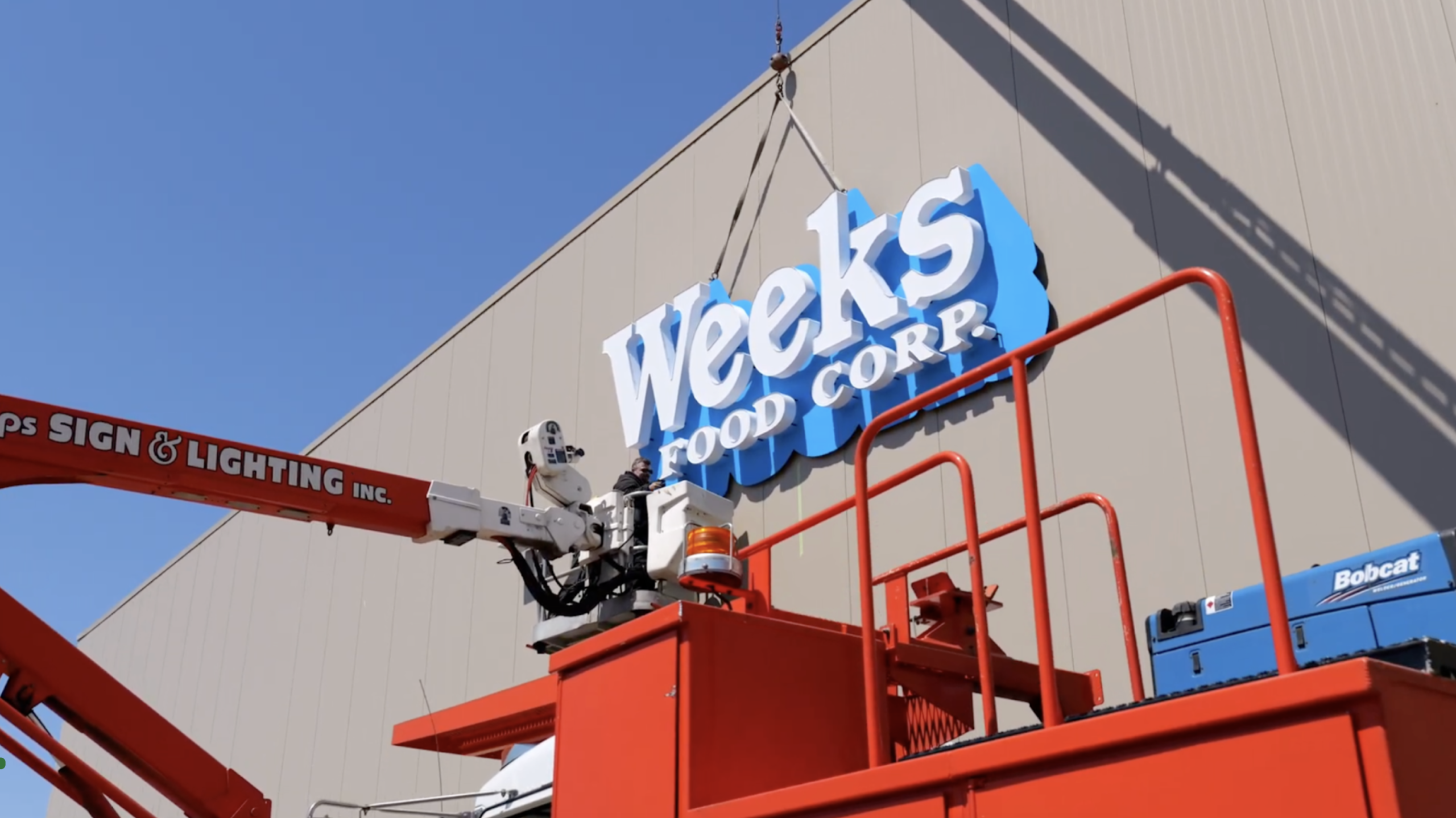 Close-up of the Weeks Food Corp. sign being installed by a crane operator, showing the bright blue background of the sign against the light gray building.